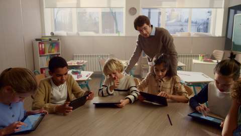 Six young students with tablets sit at a shared classroom table, and a teacher looks over one student’s shoulder at in-progress work